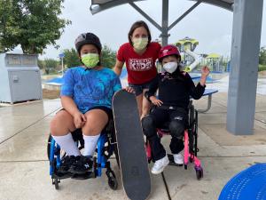 Two children in wheelchairs from Kinetic Kids pose with a skateboard and a Kinetic Kids organizational representative