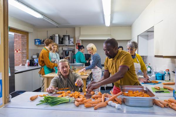 A diverse group of people work together in a commercial kitchen; in the foreground two people dice carrots and green onions to add to a baking dish. 