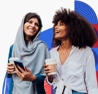 Dos mujeres caminan juntas y sonríen mientras sostienen vasos con café. Una mujer usa una túnica que cubre su cabeza mientras la otra mujer tiene pelo rizado de color oscuro.