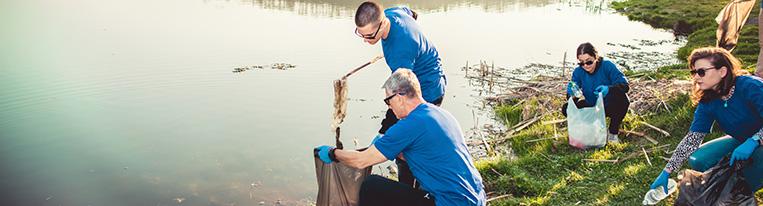 Volunteers wearing gloves and holding trash bags clean up litter at a lake.