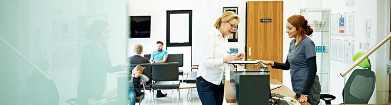 A woman stands at a desk and fills out paperwork in a health care provider's waiting room.