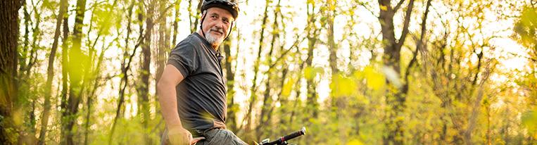 A man wearing a bicycle helmet and biking on a wooded trail pauses to look over his shoulder.