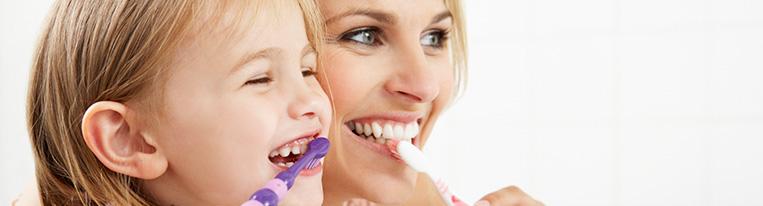 A woman and her young daughter smile while brushing their teeth over a sink.